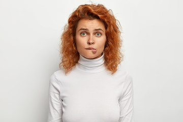 Wall Mural - Curly haired ginger female bites lips, has nervous troubled expression, hesitates about something, being sad and embarrassed, wears white jumper, looks straightly at camera. Human emotions, feelings