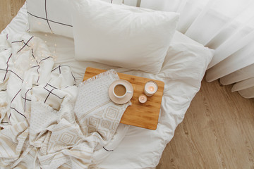 Wooden tray of coffee and candles on bed. White bedding sheets with striped blanket and pillow. Breakfast in bed. Hygge concept.