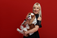 Portrait Of Smiling Young Blond Woman In Braces And Stylish Black Dress Embracing King Charles Spaniel Dog. Owner And Pet Relations Concept. Veterinary Health. Isolated Front View On Red Background.