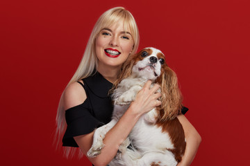 Wall Mural - Portrait of smiling young blond woman in braces and stylish black dress embracing king Charles spaniel dog. owner and pet relations concept. Veterinary health. Isolated front view on red background.