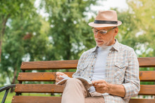 Senior Man In Hat Reading Book While Sitting On Public Bench In Park Zone