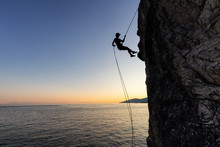 Silhouette Of A Unrecognizable Man Rappelling Down A Steep Cliff On The Rocly Ocean Coast During A Sunny Summer Sunset. Taken In Lighthouse Park, West Vancouver, British Columbia, Canada.