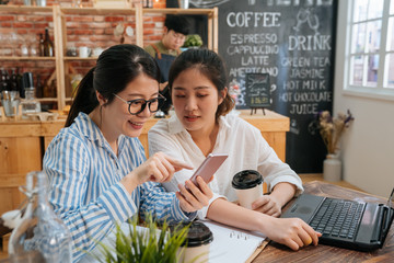 Wall Mural - Business team partners working on laptop computer at coffee bar. two young girls colleagues looking at mobile phone together in modern cafe store. barista in apron prepare customer order in counter