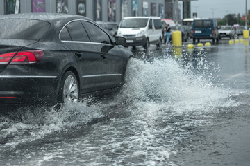  Driving car on flooded road during flood caused by torrential rains. Cars float on water, flooding streets. Splash on the car. Flooded city road with a large puddle