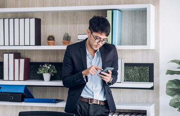 Wall Mural - Young Asian businessman wear suit take a break and using mobile for social media when standing at desk table in modern office.digital lifestyle.