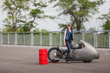 Fashion sexy woman is standing near barrel of oil with classical race motorbike. Girl biker with perfect fit slim body on highway. The spirit of freedom and independence. 