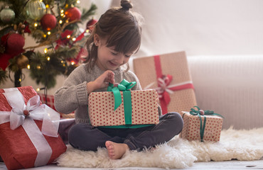 Pretty little girl is holding a gift box and smiling while sitting on her bed in her room at home