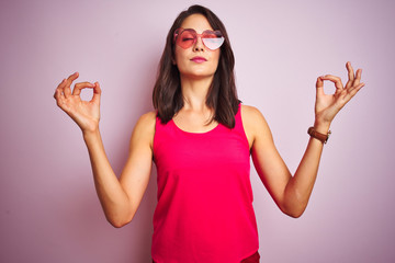 Wall Mural - Beautiful woman wearing funny pink heart shaped sunglasses over pink isolated background relax and smiling with eyes closed doing meditation gesture with fingers. Yoga concept.