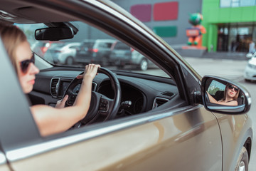 A girl driving a right-hand drive car is parked in a parking lot, near a shopping center, left-hand traffic. Close-up rear view mirror, steering wheel driver with glasses.