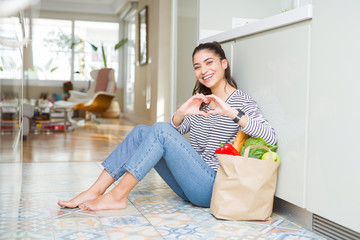 Wall Mural - Young woman sitting on the kitchen floor with a paper bag full of fresh groceries smiling in love showing heart symbol and shape with hands. Romantic concept.