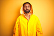 © Krakenimages.com - Young indian man wearing raincoat with hood standing over isolated yellow background with serious expression on face. Simple and natural looking at the camera.