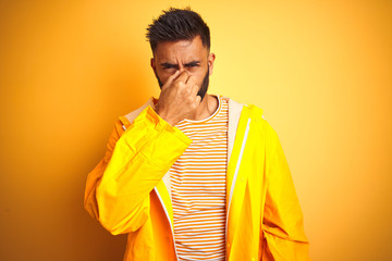 Canvas Print - Young indian man wearing raincoat standing over isolated yellow background smelling something stinky and disgusting, intolerable smell, holding breath with fingers on nose. Bad smells concept.