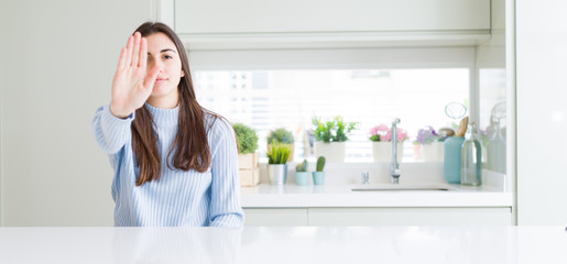 Poster - Wide angle picture of beautiful young woman sitting on white table at home doing stop sing with palm of the hand. Warning expression with negative and serious gesture on the face.