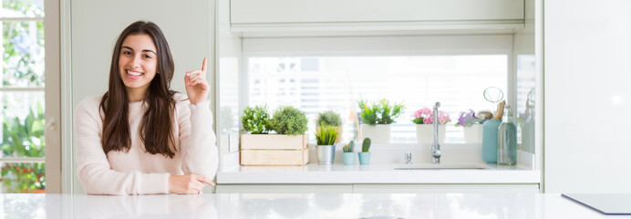 Wide angle picture of beautiful young woman sitting on white table at home with a big smile on face, pointing with hand and finger to the side looking at the camera.