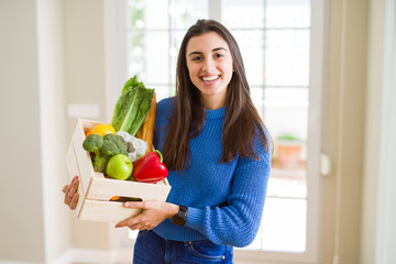 Canvas Print - Beautiful young woman holding wooden box full of healthy groceries with a happy face standing and smiling with a confident smile showing teeth