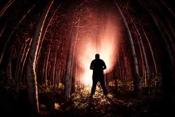 strange light in a dark forest at night. Silhouette of person standing in the dark forest with light. Dark night in forest at fog time. Surreal night forest scene.