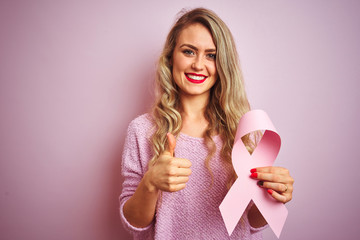 Young beautiful woman holding cancer ribbon over pink isolated background happy with big smile doing ok sign, thumb up with fingers, excellent sign