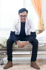 Portrait of Asian young male doctor with white lab coat sitting on indoors hospital clinic bed looking at camera