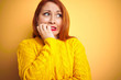 © Krakenimages.com - Young redhead woman wearing winter sweater standing over isolated yellow background looking stressed and nervous with hands on mouth biting nails. Anxiety problem.