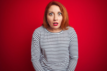 Wall Mural - Young redhead woman wearing strapes navy shirt standing over red isolated background afraid and shocked with surprise expression, fear and excited face.