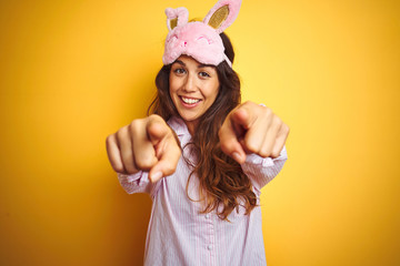 Young woman wearing pajama and sleep mask standing over yellow isolated background pointing to you and the camera with fingers, smiling positive and cheerful