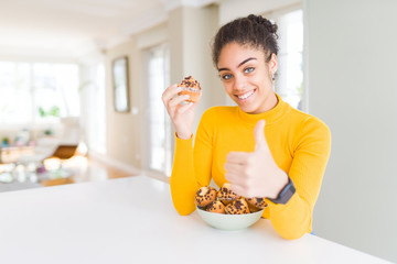 Young african american woman eating chocolate chips muffins happy with big smile doing ok sign, thumb up with fingers, excellent sign