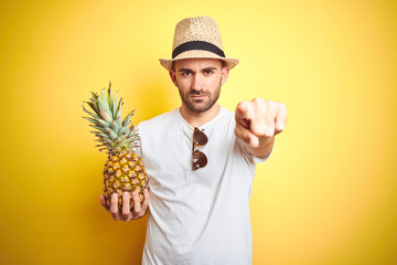 Wall Mural - Young man wearing summer hat and holding pineapple over yellow background pointing with finger to the camera and to you, hand sign, positive and confident gesture from the front