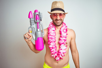 Poster - Young man wearing summer hat and hawaiian lei flowers holding water gun over isolated background with a happy face standing and smiling with a confident smile showing teeth