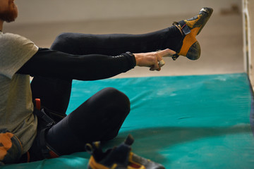 Close up view on strong legs, unrecognizable male climber with beard puts on climbing shoes, getting ready to intensive bouldering training, sitting on blue crash pad. Cropped shot.