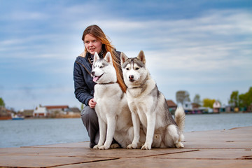 Young beautiful girl with red hair is sitting on a pier with two husky dogs. Woman with a pair of Siberian Husky