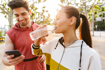 Sticker - Photo of sporty pleased man and woman using cellphone and drinking water while working out in city boulevard