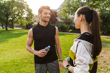 Canvas Print - Photo of young nice man and woman talking together and holding plastic bottle while working out in city park