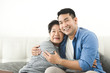 © nuiiko - Asian mother hugging her son and sitting on sofa at home, lifestyle concept.