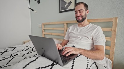 Poster - Happy bearded man using laptop computer while lying on bed in bedroom