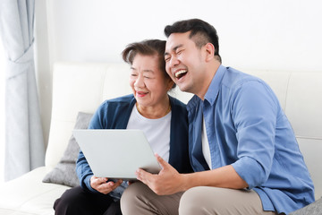 Wall Mural - Asian young man and senior woman using laptop computer in living room concept Son teach mother use computer laptop