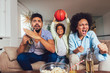 © Mediteraneo - Happy African American family of three watching tv and cheering sport games on sofa at home