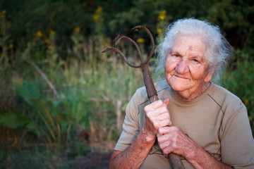 Close-up portrait of an old woman with gray hair holding a rusty pitchfork or chopper in her hands, face in deep wrinkles, selective focus