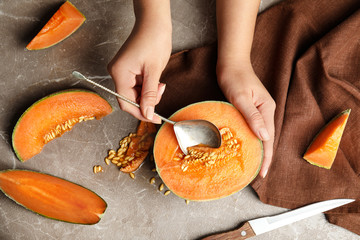 Young woman removing seeds from ripe melon with spoon at beige marble table, top view