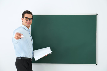 Wall Mural - Young teacher with book near blank chalkboard in classroom. Space for text