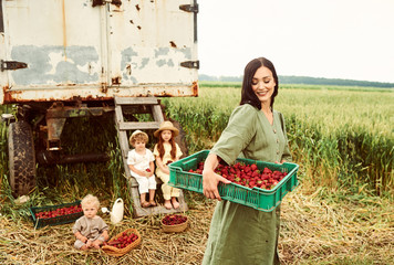 Wall Mural - Beautiful young caucasian mother with her children in a linen dress with a basket of strawberries gathers a new crop and has fun with the children near the trailer