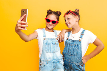 Wall Mural - Two teenage girls, girlfriends have a rest and take a selfie on a smartphone on a yellow background