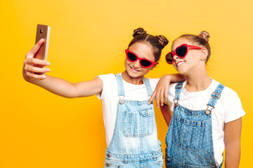 Wall Mural - Two teenage girls, girlfriends have a rest and take a selfie on a smartphone on a yellow background