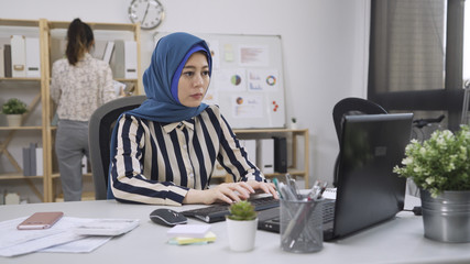 Canvas Print - muslim woman connected on internet with laptop typing on keyboard replying emails to client order. japanese colleague standing in background gind document files in bright cozy office. multi ethnic.