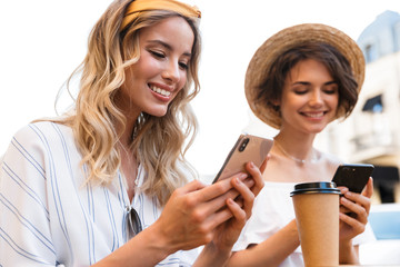 Canvas Print - Cheerful young pleased optimistic girls friends sitting outdoors in cafe drinking coffee using mobile phones.