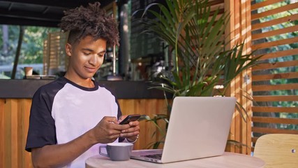 Canvas Print - Happy african student writing message on smartphone while sitting by the table with laptop computer in cafe outdoors