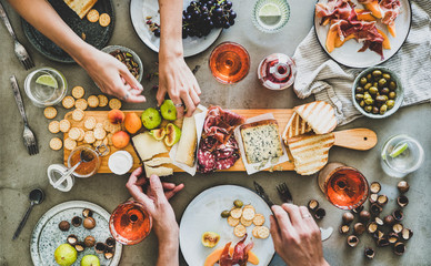 Wall Mural - Mid-summer picnic with wine and snacks. Flat-lay of charcuterie and cheese board, rose wine, nuts, olives and peoples hands over concrete table background, top view. Family, friends holiday gathering