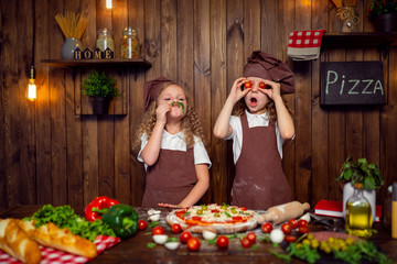 Adorable little girls chef wearing aprons and headbands cooking pizza and making face with tomatoes instead eyes and lettuce, opened mouth on stylish wooden kitchen