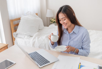 Poster - Asian businesswoman drinking coffee and while working with laptop on table in bedroom at home.Work at home concept.work from home concept