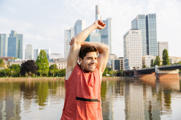 Canvas Print - Photo of smiling athletic man stretching arm while working out near city riverfront