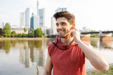 Canvas Print - Photo of smiling athletic man using earphones while working out near city riverfront